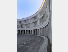 Edificio monumentale a pianta semicircolare con facciata in pietra chiara, colonnato al piano terra e lunghe file di finestre verticali ai piani superiori. Davanti si apre una grande area pavimentata curva, con marciapiede e bandiere esposte sotto il portico.