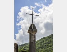 Croce metallica montata sulla sommità di una colonna in pietra all’aperto, ripresa contro un cielo azzurro con nuvole. Sullo sfondo si vede un pendio montano coperto di vegetazione verde e un angolo di edificio in pietra.