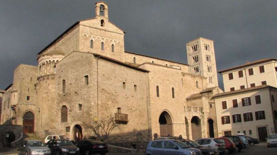 Grande chiesa in pietra in stile romanico con facciata massiccia, portali ad arco e piccole finestre, affiancata da un campanile quadrato. L’edificio si affaccia su una piazza con auto parcheggiate e palazzi adiacenti sotto un cielo scuro.