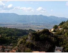 Veduta panoramica da una collina con vegetazione e rocce, con alcune case e una strada sul versante. Sullo sfondo si estende una pianura con campi e corsi d’acqua, chiusa da una catena montuosa sotto un cielo parzialmente nuvoloso.