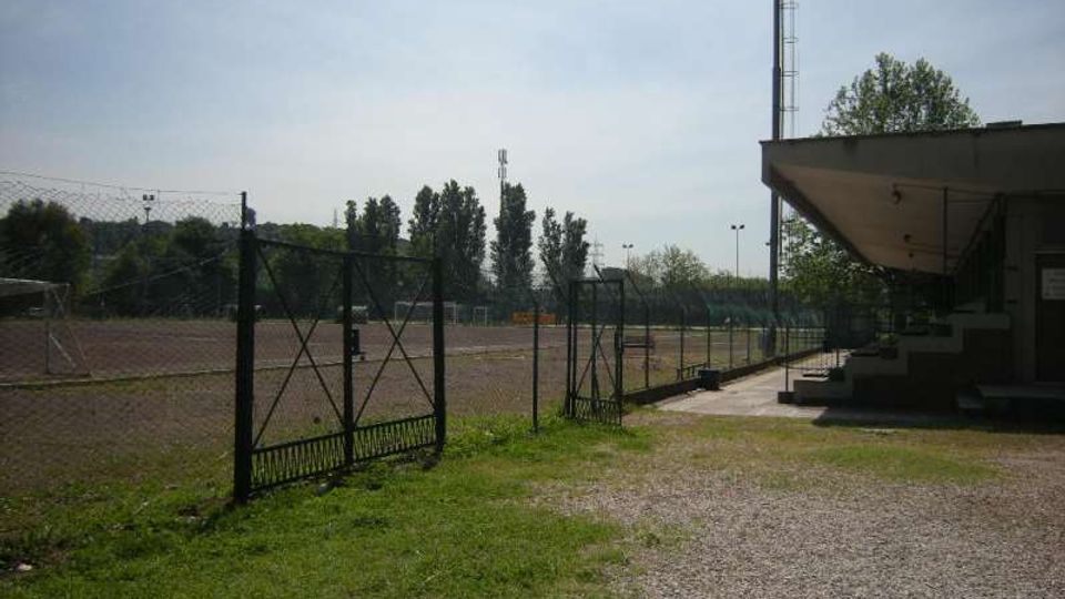 Campo sportivo all’aperto recintato con rete metallica e cancello, affiancato da un vialetto in ghiaia e da una piccola tribuna coperta sulla destra. Sullo sfondo si vedono alberi e alti pali per l’illuminazione sotto un cielo sereno.