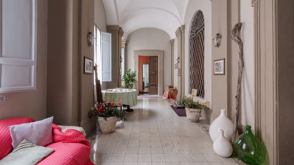 Corridoio interno di un edificio storico con soffitto a volte, colonne e pavimento piastrellato chiaro. Lungo le pareti ci sono finestre alte, lanterne, quadri e decorazioni con vasi, piante, tappeti e una zona divano rossa; in fondo si vede una porta aperta.