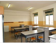 Aula laboratorio con banchi di lavoro in metallo e sgabelli, pavimento in cemento e pareti bianche con fascia gialla. Due finestre con avvolgibili e lampade fluorescenti a soffitto illuminano lo spazio.