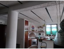 Cucina abitabile con soffitto a travi scure a vista, pensili in legno e piano cucina angolare con forno e piastrelle decorative. Una finestra con vetri verde-azzurri illumina la zona lavabo, con lampada a sospensione sopra il bancone.
