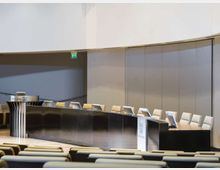 Sala conferenze moderna con un lungo banco curvo in metallo e legno, dotato di microfoni e schermi, e file di sedute in platea in primo piano. Sullo sfondo si vedono pareti lisce e pannelli chiari con un’uscita di emergenza segnalata.