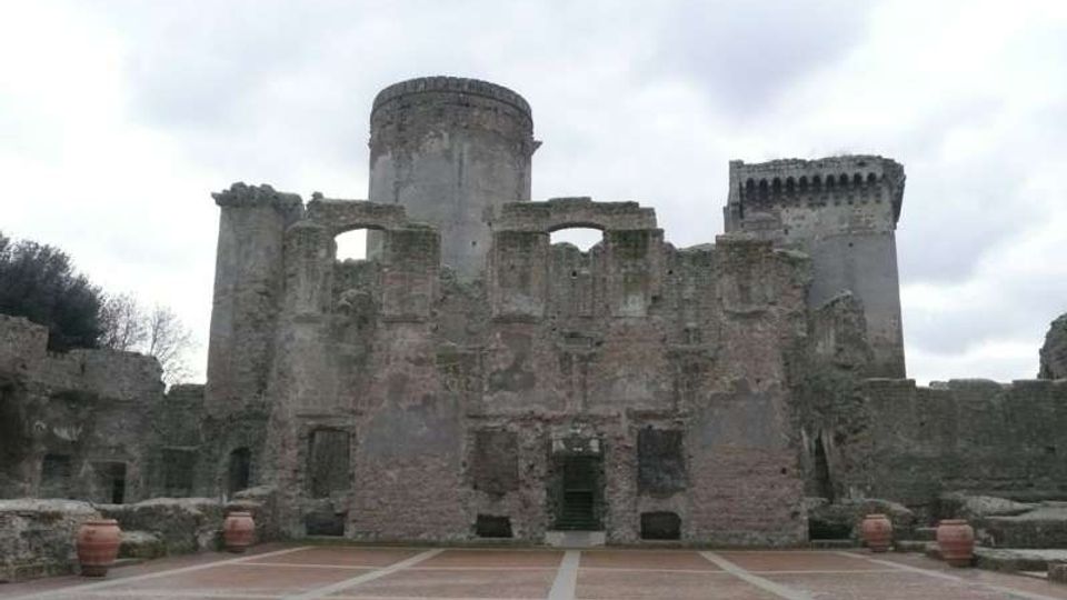Rovine di un castello in pietra con muri parzialmente crollati e grandi aperture ad arco, affiancato da due torri cilindriche sullo sfondo. In primo piano si vede un ampio piazzale pavimentato con linee geometriche e alcuni vasi in terracotta ai lati.