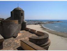 Veduta costiera con una fortificazione in muratura e torre semicircolare in primo piano, affacciata su una lunga spiaggia sabbiosa. Sullo sfondo si vedono il mare, un porto con numerose barche e un lungo molo frangiflutti.