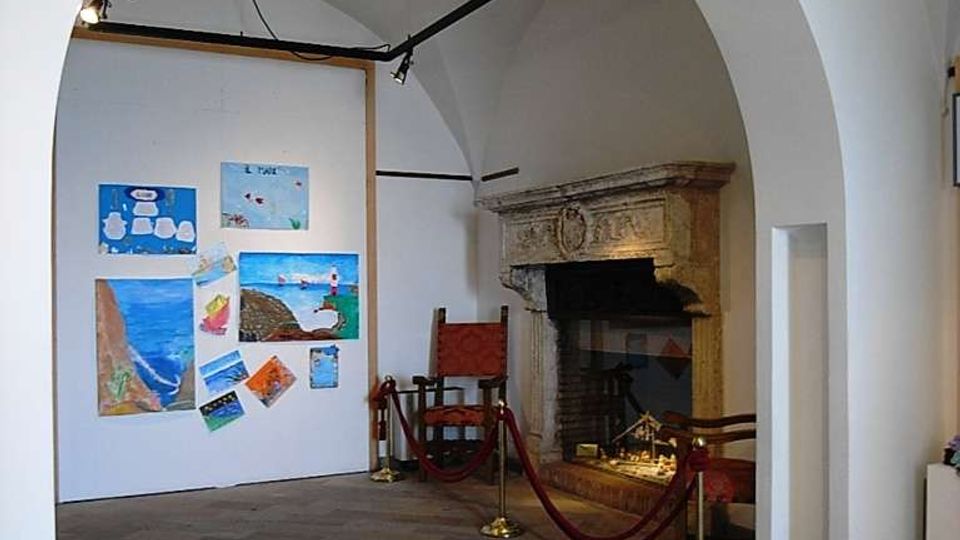 Interno di una sala espositiva con soffitto a volte e grande arco bianco, pavimento in pietra e illuminazione a binari. Sulla sinistra c’è una parete con disegni e dipinti appesi, mentre sulla destra si vede un grande camino in pietra delimitato da cordoni rossi.