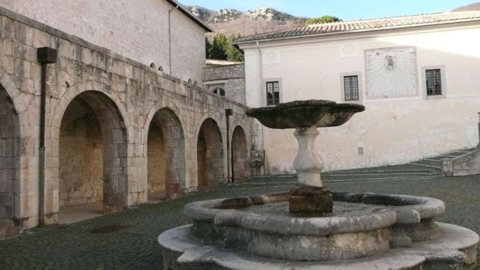 Cortile lastricato con una grande fontana in pietra al centro, composta da una vasca circolare e una coppa superiore su piedistallo. Sullo sfondo si vedono un porticato in pietra con archi e un edificio intonacato chiaro con finestre e una meridiana, con colline rocciose sullo sfondo.