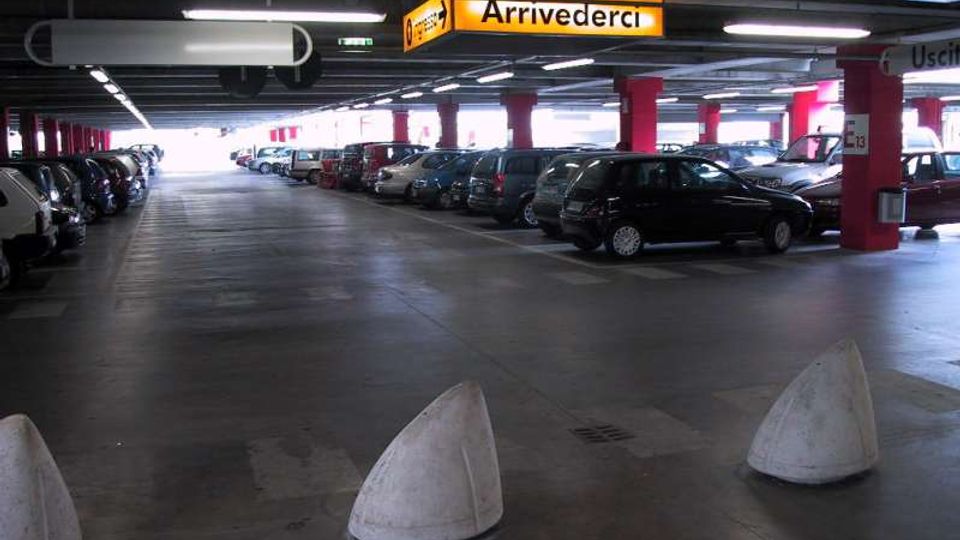 Parcheggio multipiano coperto con file di auto parcheggiate ai lati, colonne rosse e soffitto basso con illuminazione a neon. In alto sono visibili cartelli gialli di segnaletica, tra cui “Arrivederci” e “Uscita”.