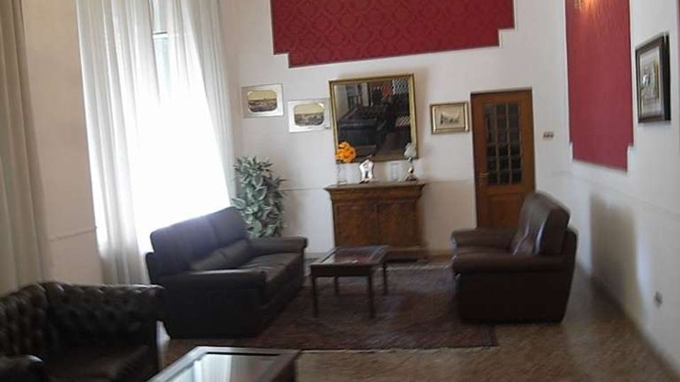 Ampio salone con soffitto alto, lampadario centrale, grandi finestre con tende chiare e pareti bianche con riquadri decorativi rossi. L’ambiente è arredato con divani e poltrone in pelle scura, tavolini, tappeto e una credenza con specchio e quadri alle pareti.