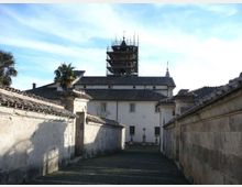 Vicolo in discesa fiancheggiato da alti muri intonacati con tetti in coppi, che conduce a un edificio bianco in stile tradizionale. Sullo sfondo si vede una torre con impalcature di restauro e una piccola cupola, sotto un cielo azzurro.