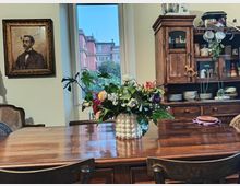 Sala da pranzo domestica con grande tavolo in legno lucido e vaso di fiori al centro, affiancata da un’ampia finestra che dà su palazzi e alberi. Sullo sfondo si vede una credenza in legno con piatti e tazze a vista e, sulla parete, un quadro incorniciato.