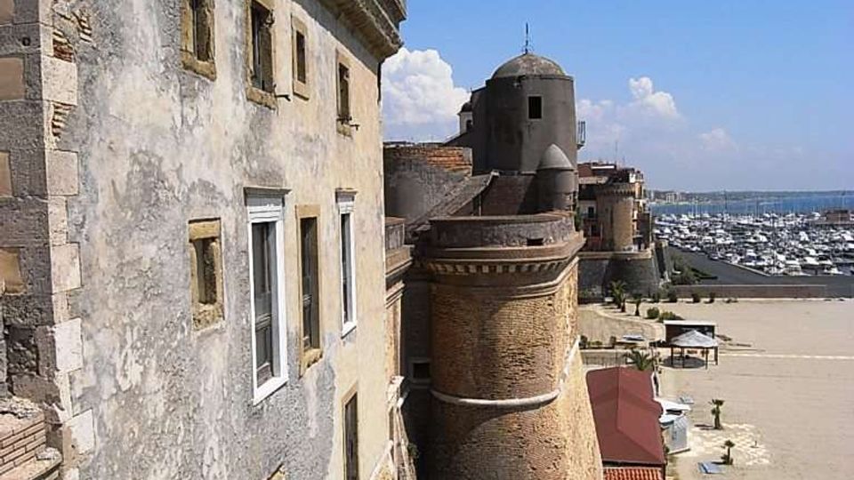 Veduta laterale di una fortezza costiera in muratura, con torre cilindrica e mura in pietra e intonaco, affacciata su un’area aperta vicino al mare. Sullo sfondo si vede un porto turistico con numerose barche e il litorale sotto un cielo sereno.
