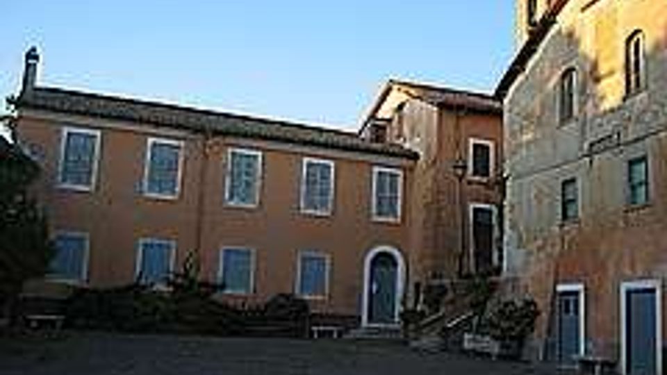 Cortile lastricato con panchine e alcune piante, circondato da edifici in muratura color ocra e mattoni con finestre regolari. Sullo sfondo si vedono porte ad arco e facciate storiche illuminate dalla luce bassa, sotto un cielo sereno.