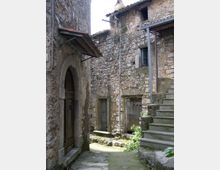 Vicolo stretto in un borgo con edifici in pietra grezza, porte ad arco e piccole finestre, con cavi e tubazioni a vista sulle facciate. A destra una scalinata in pietra sale verso un pianerottolo coperto da una tettoia metallica.