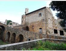 Edificio storico in pietra con facciate irregolari e piccole finestre, affiancato da un lungo muro con arcate in muratura e una rampa in salita. Sullo sfondo si vedono resti di strutture in pietra e vegetazione in un contesto rurale all’aperto.
