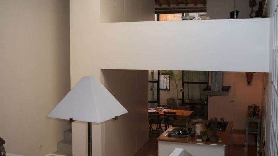 Interno di una casa moderna su più livelli con soppalco e parapetti bianchi, soffitto con travi in legno a vista e pavimento in legno. In basso si vede una cucina open space con isola e zona pranzo sullo sfondo, illuminata da lampade con paralume bianco.