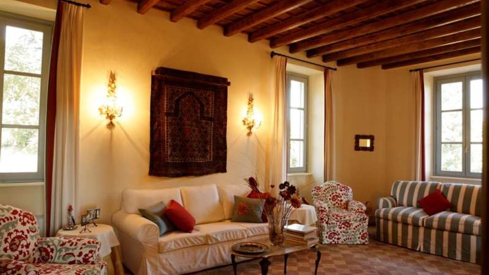 Soggiorno in stile rustico con soffitto a travi in legno a vista, pareti chiare e pavimento a piastrelle. La stanza è arredata con divani e poltrone imbottite, tavolino centrale e grandi finestre con tende, illuminate da applique a parete.
