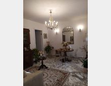 Sala da pranzo in stile classico con pavimento in marmo, grande lampadario di cristallo e pareti bianche con applique. Al centro c’è un tavolo rotondo con sedie trasparenti, specchio decorativo alla parete e mobili in legno con tappeti e piante ornamentali.