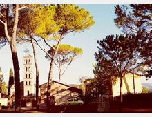 Veduta di un complesso storico con campanile in pietra a più ordini di bifore, affiancato da edifici bassi con tetti in coppi. In primo piano un’area alberata con alti pini mediterranei che incorniciano la scena.