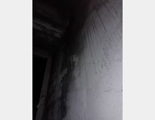 Parete interna di un corridoio o vano stretto, con intonaco grigio sporco e colature evidenti di umidità. In alto si vede il soffitto e a sinistra un tubo, con un’area in ombra che chiude lo spazio.