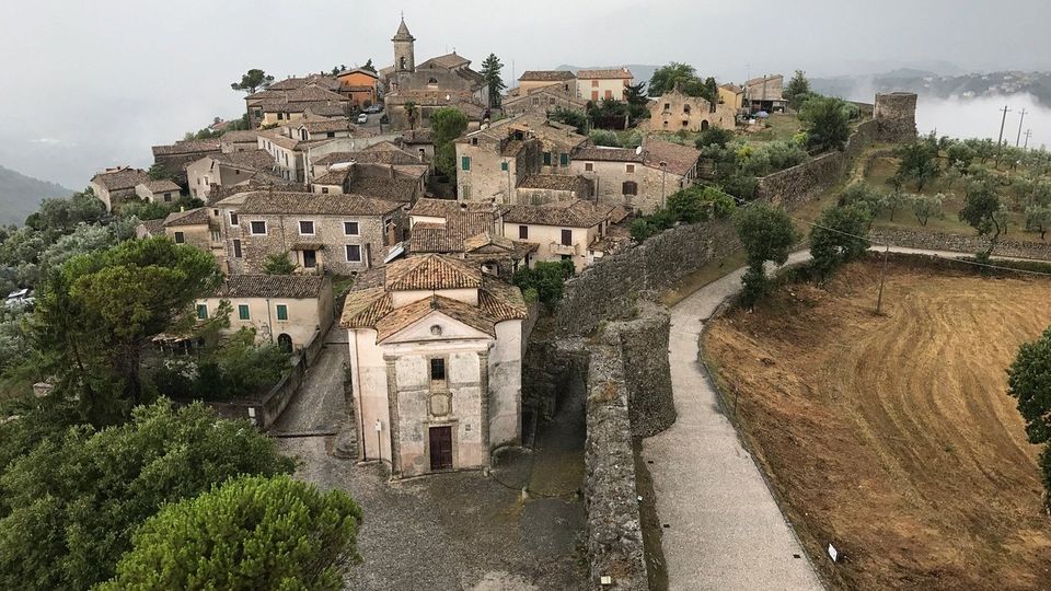 Veduta dall’alto di un borgo collinare con case in pietra e tetti in coppi, una chiesa in primo piano e un campanile al centro. Un muro di cinta in pietra segue una strada sterrata ai margini, con campi e uliveti circostanti sotto un cielo nuvoloso.