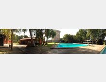Area esterna con piscina rettangolare e solarium in pietra, circondata da alberi. Sullo sfondo si vedono un edificio con tetto in tegole rosse e una torre in pietra, in un contesto verde e soleggiato.