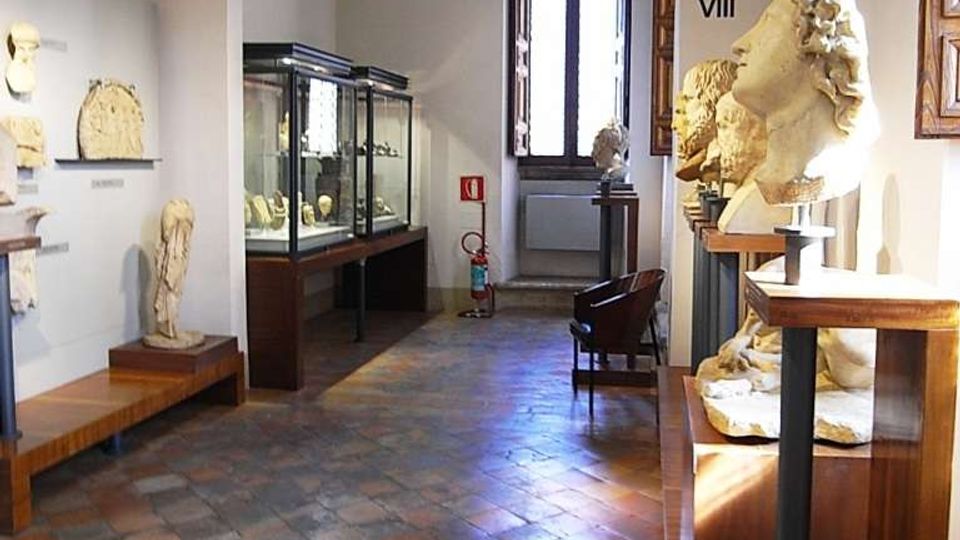 Interno di una sala museale con pavimento in cotto e soffitto scuro, attraversata da un grande arco che conduce a una finestra sul fondo. Lungo le pareti e su tavoli espositivi ci sono busti e frammenti scultorei, con vetrine in vetro illuminate da faretti.