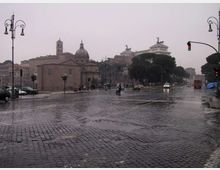 Ampia strada cittadina lastricata in sampietrini bagnati dalla pioggia, con lampioni e semafori lungo i lati e traffico sparso. Sullo sfondo si vedono edifici storici con cupole e campanile, alberi ad alto fusto e un grande monumento su un’altura.