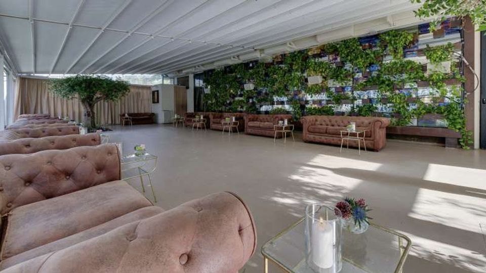 Ampio salone coperto con soffitto bianco a pannelli e pavimento chiaro, arredato con divani capitonné color cipria e tavolini in vetro con candele. Sul fondo si vede una grande parete verde verticale con piante e pannelli decorativi, affiancata da tende e un albero ornamentale.