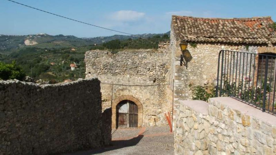 Stradina in pietra in discesa tra muri in pietra e case rustiche con tetto in coppi, con un portale ad arco in mattoni e una lanterna a parete. Sullo sfondo si vedono colline verdi e campagna sotto un cielo azzurro.