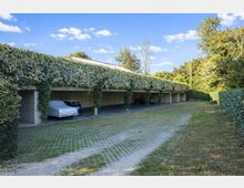 Parcheggio coperto all’aperto con una lunga tettoia in cemento sostenuta da pilastri, ricoperta di edera e con posti auto in ombra. Davanti c’è un vialetto pavimentato con griglie erbose, circondato da siepi e alberi sotto un cielo azzurro.