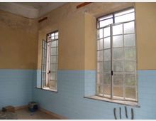 Interno di una stanza spoglia con pareti bicolore (giallo sopra e piastrelle azzurre sotto) e due grandi finestre a riquadri con vetro opaco, una delle quali aperta. Il soffitto è alto e le finestre danno su un ambiente esterno indistinto.
