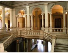 Interno di un edificio storico con cortile a doppia altezza, caratterizzato da colonne e arcate in pietra. Una scala monumentale con balaustre conduce a un ballatoio superiore affacciato sull’atrio con pavimento lucido e grandi porte.