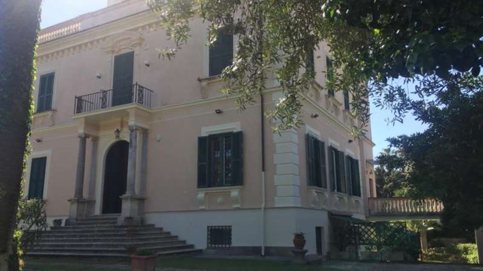 Villa storica a due piani con facciata chiara, persiane scure e ingresso con scalinata e portico a colonne. Intorno si vede un giardino con vialetto in pietra e alberi che incorniciano l’edificio, con una balaustra sul lato destro.