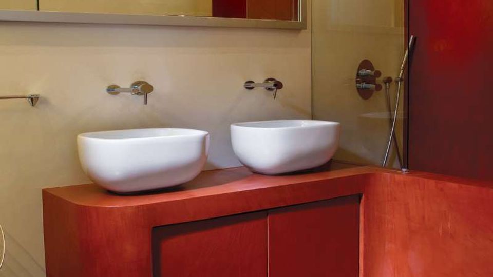 Bagno moderno con pavimento e rivestimenti in resina rosso intenso, mobile lavabo coordinato con due lavabi a bacinella e rubinetteria a parete sotto uno specchio lungo. A destra è visibile una doccia con parete in vetro e soffione, con un sanitario bianco in primo piano.