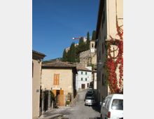 Strada stretta in un borgo con case in pietra e facciate chiare, tetti in coppi e auto parcheggiate lungo il margine. Sullo sfondo si vede una fortificazione su una collina con cipressi e una torre con orologio, sotto un cielo sereno.