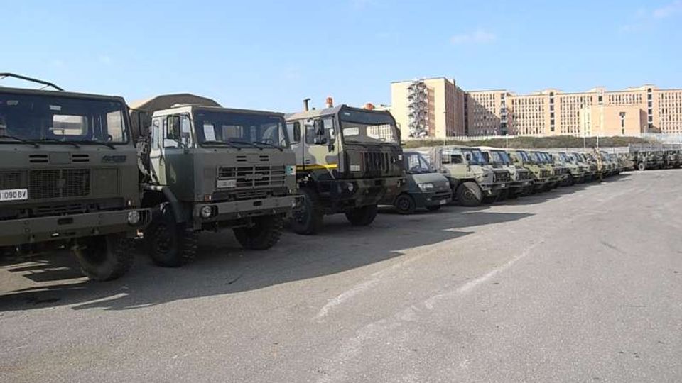 Parcheggio asfaltato all’aperto con una lunga fila di camion e veicoli militari verde oliva allineati. Sullo sfondo si vedono grandi edifici in cemento e mattoni sotto un cielo sereno.