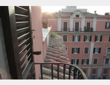 Vista da un piccolo balcone con ringhiera metallica e persiana in legno, affacciato su tetti in tegole e su un edificio urbano rosa a più piani con finestre e persiane scure. Sullo sfondo si vedono altri tetti e una luce di controluce.