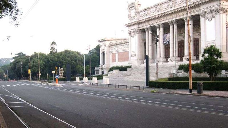 Ampio viale urbano con binari del tram in primo piano, affiancato da alberi e arredi stradali. Sulla destra si vede un grande edificio monumentale in stile neoclassico con facciata in pietra, colonne alte, portico e scalinata d’ingresso.