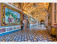 Lungo corridoio interno di un palazzo storico, con pavimento a mosaico geometrico in marmo e pareti rivestite da grandi affreschi incorniciati e decorazioni policrome. Il soffitto a volta è riccamente ornato con dipinti e stucchi, illuminato da lampadari, con porte in pietra e statue lungo il percorso.
