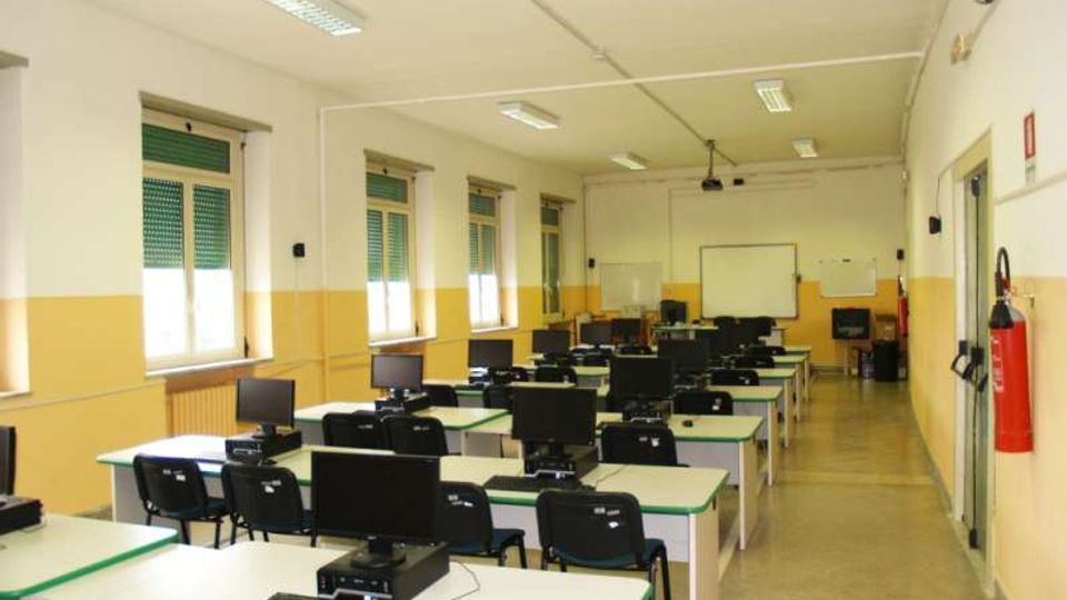 Aula informatica con file di banchi dotati di computer e monitor, pareti bianche e gialle e illuminazione a neon sul soffitto. Lungo un lato ci sono finestre con tapparelle verdi e, in fondo, una lavagna con proiettore a soffitto.