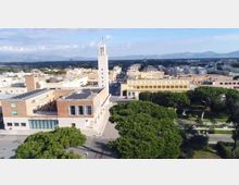 Veduta aerea di un complesso civico moderno con torre bianca centrale e edifici bassi in mattoni e intonaco, affacciati su una piazza. In primo piano un parco con alberi e vialetti, sullo sfondo la città e una catena di montagne.
