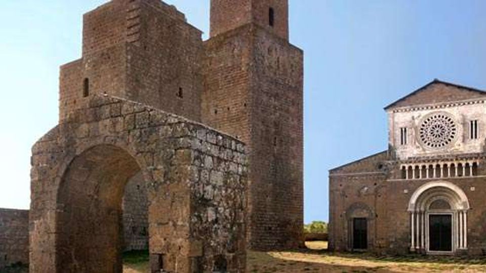 Complesso medievale in pietra con grande arco d’ingresso e massicce torri quadrangolari, accanto a una chiesa romanica con facciata a capanna, rosone centrale e portale ad arco. L’insieme è in un’area aperta con terreno erboso e cielo sereno sullo sfondo.