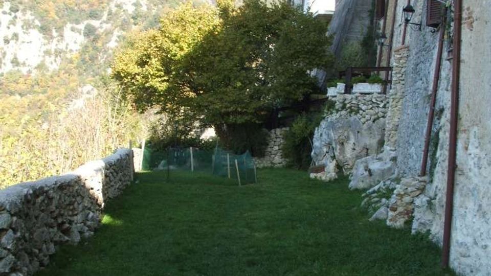 Striscia di prato lungo un edificio in pietra su un pendio, con muretto a secco sul lato sinistro e rocce affioranti vicino alla facciata. Sullo sfondo si vedono colline boscose e montagne.