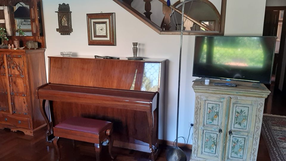 Soggiorno arredato con pianoforte verticale in legno e sgabello, accanto a un mobile dipinto con TV sopra e una lampada da terra. Sullo sfondo si vedono un grande armadio intagliato, quadri alle pareti e una scala in legno con parapetto decorato.