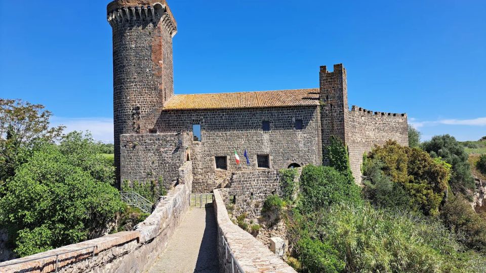 Castello medievale in pietra con alta torre cilindrica merlata e mura fortificate, visto da un lungo camminamento sopraelevato che conduce all’ingresso. Intorno si estende una vegetazione fitta in campagna sotto un cielo azzurro limpido.