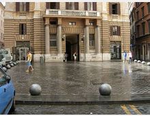 Facciata di un edificio storico in pietra con portico sorretto da colonne e grande ingresso ad arco, affacciato su una piazza pavimentata in sampietrini. In primo piano si vede una fila di dissuasori sferici in pietra lungo il bordo del marciapiede.