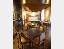 Sala interna tipo lounge o sala da tè con tavoli e sedie in legno, pavimento in marmo e soffitto con pannelli in legno. Sul fondo ci sono scaffali con libri e un tavolo da biliardo coperto, con una grande finestra laterale che illumina l’ambiente.